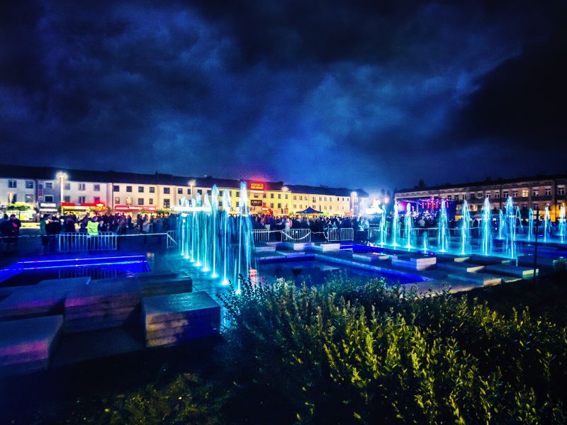 Zdjęcie pl. Kościuszki, wykonanane nocą. Granatowe niebo, widoczne podświetlone na niebiesko fontanny i w tle sylwetki ludzi