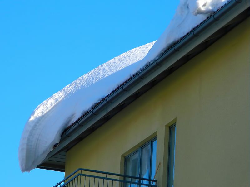 Na zdjęciu budynek mieszkalny z dachem, na którym wisi zaspa śnieżna. Budynek w kolorze żółtym z fragmentem balkonu, zwisający śnieg z dachu po świeżych opadach