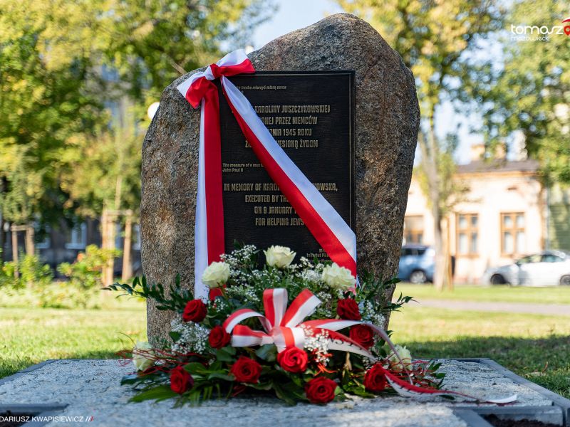 Zdjęcie obelisku z tablicą pamiątkową, przepasany biało-czerowną szarfą, przed leży wiązanka biało-czerwonych kwiatów
