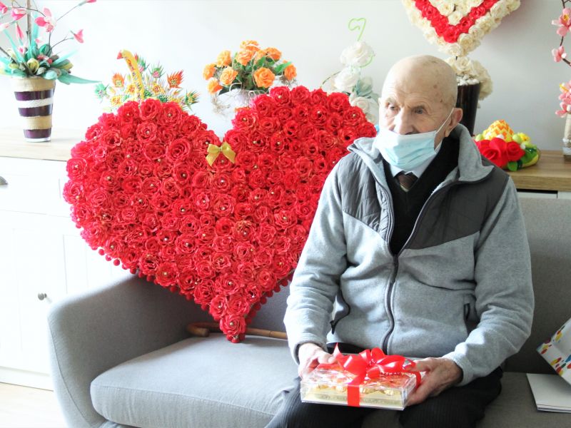  starszy mężczyzna w maseczce  siedzi na kanapie obok niego serce z bibułowych róż
