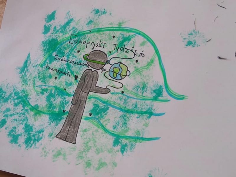 Zdjęcie przedstawia dziecięcy rysunek przedstawiający projekt logo Europejskiego Tygodnia Zrównoważonego Transportu. Na rysunku postać człowieka-drzewa, z której wyrastają zielone gałęzie