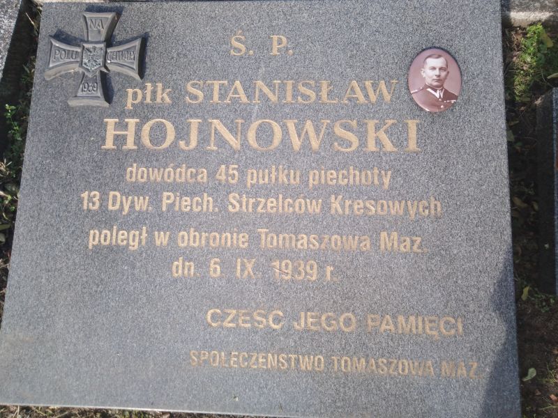 Na zdjęciu nagrobek płk Hojnowskiego, obrońcy Tomaszowa Mazowieckiego z 1939 roku. Widać płyte nagrobną, inskrypcję na nagrobku z datą urodzenia i śmierci pułkownika oraz z medalionem ze zdjęciem oficera