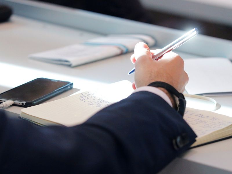 Na zdjęciu widac rękę trzymającą długopis, któa wypełnia jakieś dokumenty, zapisuje notatki w kalendarzu biurowym