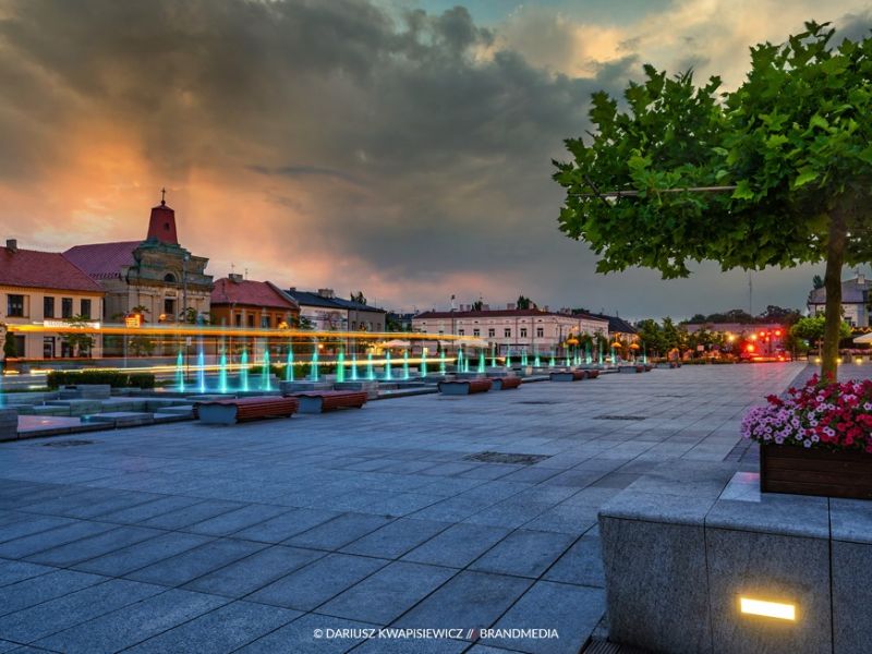 zdjęcie przedstawia plac Kościuszki wieczorem kolorowe fontanny, kwiaty w donicach, drzewo oraz dużo żółtych świateł