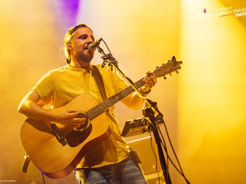 Na zdjęciu Paweł Domagała podczas koncertu w Tomaszowie Mazowieckim. Artysta w żółtej koszullce, gra na akustycznej gitarze i śpiewa do mikrofonu