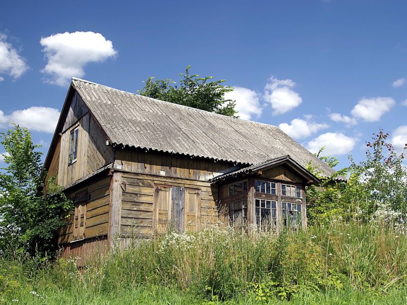 Na zdjęciu stary dom z abestowym dachem, stojący w wysokiej trawie