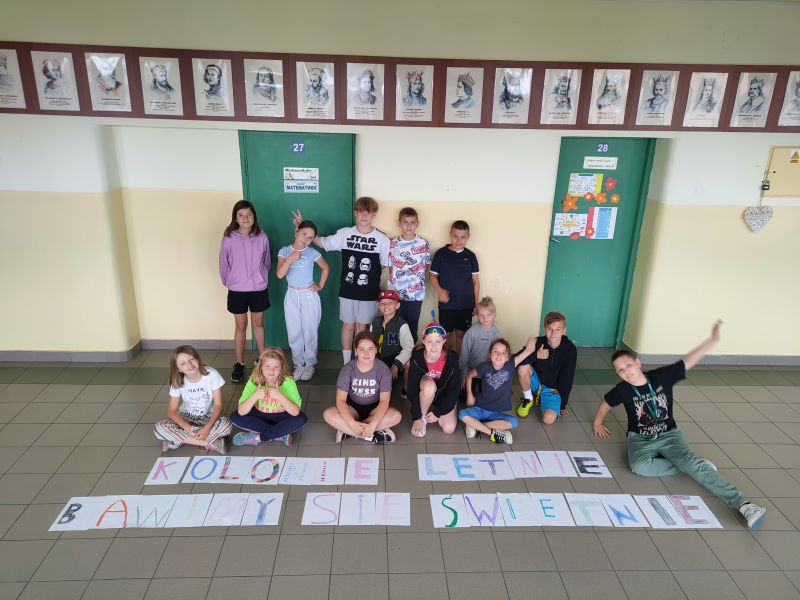 Na zdjęciu uczestnicy feriiw  Szkole Podstawowej nr 9 prezentują na korytarzu szkoplnym hasło ferii ułożone z liter zapisanych na kartkach - Kolonie Letnie - Bawimy się Świetnie