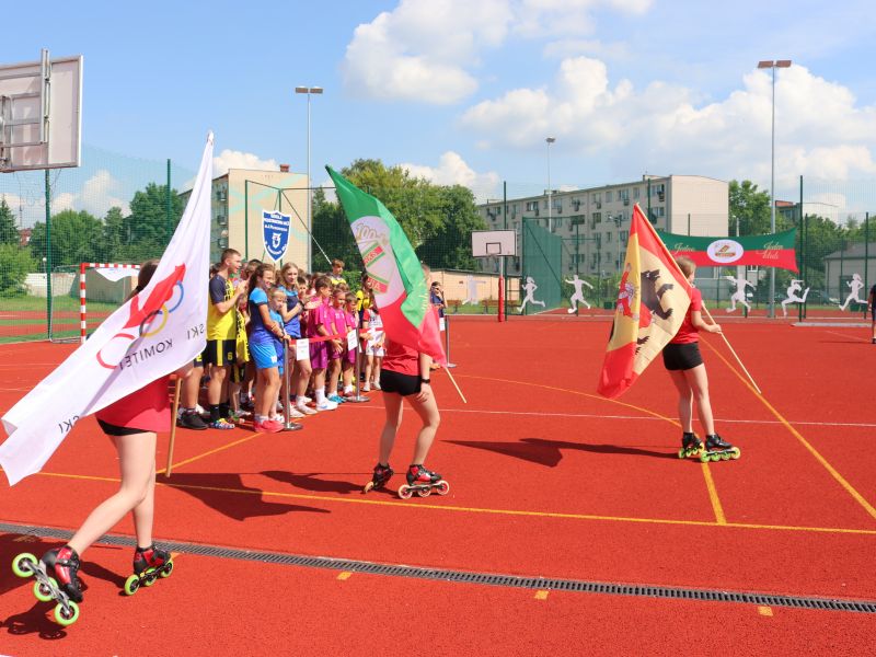Na zdjęciu dzieci z flagami i na wrotkach otwierają zawody sportowe na boisku