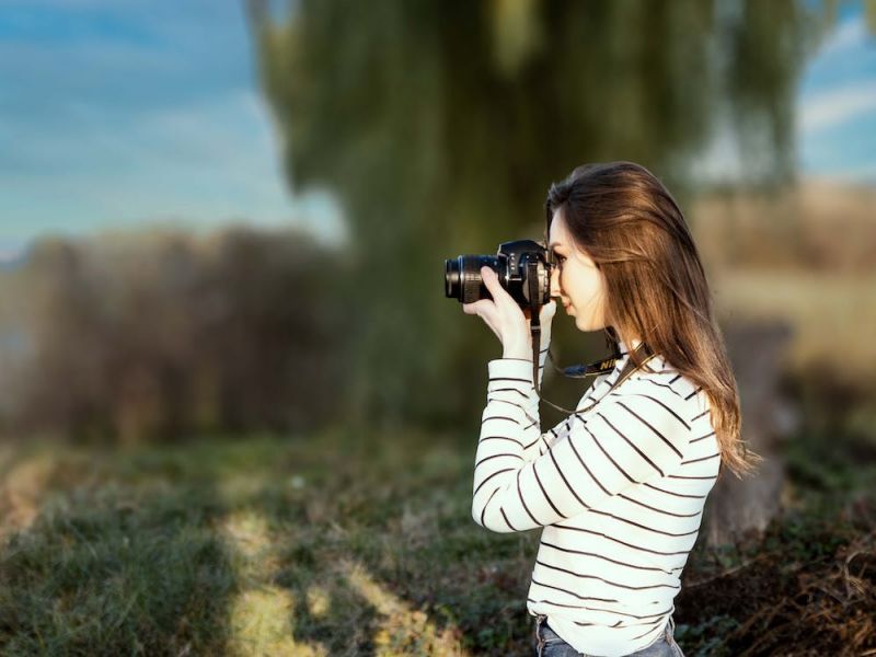 Na zdjęciu młoda dziewczyna robiąca zdjęcie aparatem fotograficznym. Dziewczyna stoi na łące