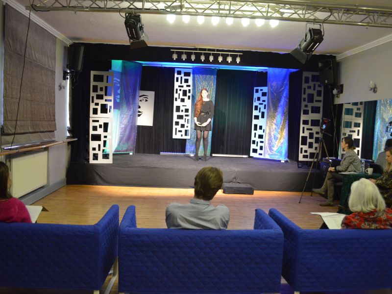 Na zdjęciu recytatorka startująca w konkursie recytatorskim Miejskiego Ośrodka Kultury. Dziewczyna recytuje tekst na scenie, występu wysłuchuje jury siedzące we fotelach