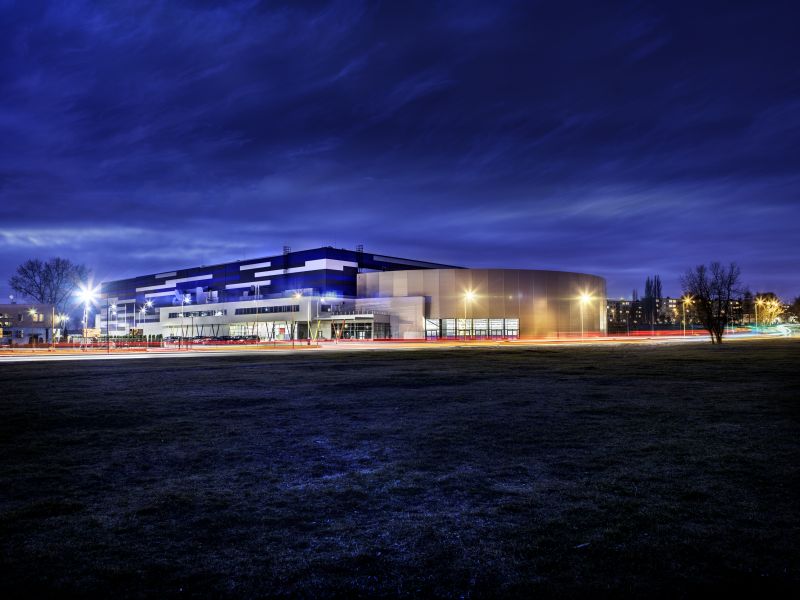 Nocne zdjęcie Areny Lodowej, granatowe niebo, ciemna trawa, w tle podświetlony, niebieski budynek Areny