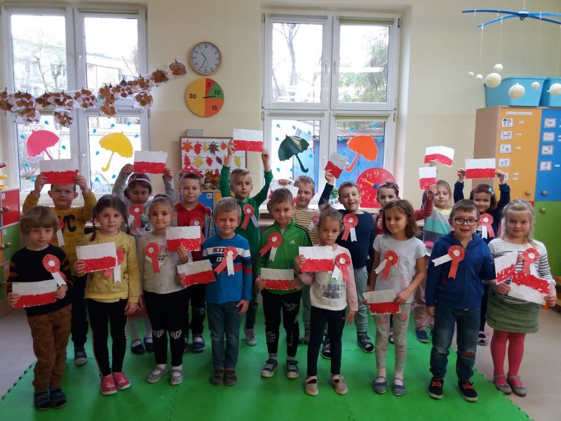 Na zdjęciu widać dzieci w wieku przedszkolnym z kotylionami biało-czerwonymi i biało-czerewonymi flagami wykonanymi na zajęciach plastycznych z okazji Święta Niepodległości. Zdjęcie wykonano w pracowni przedszkolnej.