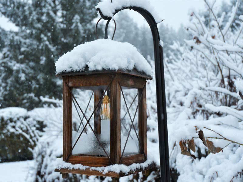 Na zdjęciu ogrodowa lampka z ośniezonym daszkiem, w tle silna zimowa aura, na gałęziach mnóstwo śniegu
