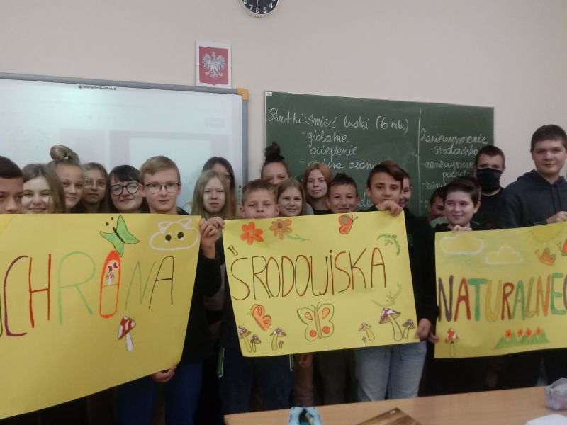 Na zdjęciu stoi grupa młodziezy szkolnej z własnoręcznie wykonanymi plakatami na temat ochrony środowiska. Zdjęcie wykonano w pracowni szkolnej, w tle tablica oraz slip-chart