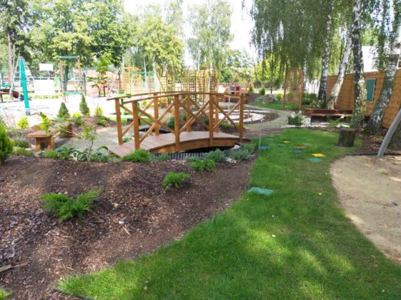 Na zdjęciu widoczny ogród sensoryczny, zielona trawa, mostek i krzewy