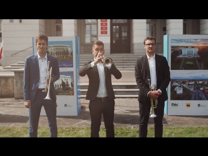 Na zdjęciu trzech muzyków, trębaczy, wykonujących hejnał Tomaszowa Mazowieckiego. W tle budynek Urzędu Miasta i widoczny fragment wystawy o historii miasta
