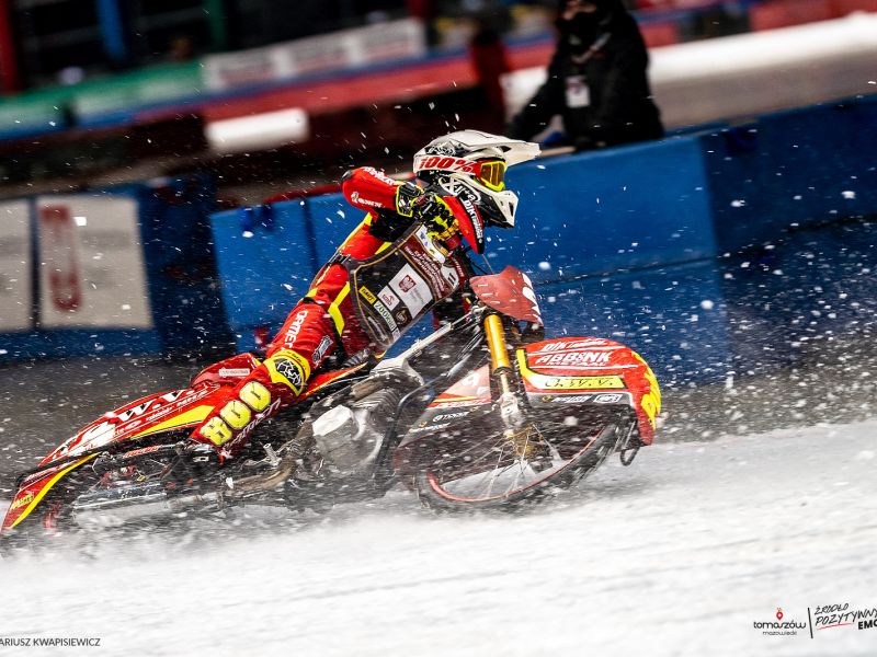 Na fotografii motocyklista podczas wyścigu na lodzie. Motocyklista ubrany w czerwony kombinezon podczas brania ostrego zakrętu. Wokół rozbryzgi lodu.