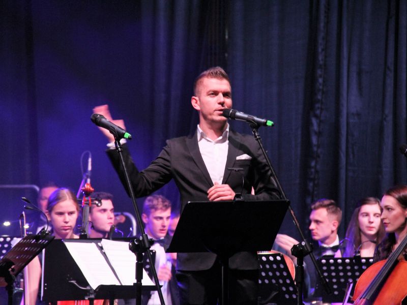 Na zdjeciu Kamil Wrona instruktor MCK, stojący na scenie przed mikrofonem. W tle muzycy Młodzieżowej Orkiestry Symfonicznej Tomaszowa Mazowieckiego
