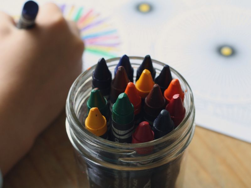 Na zdjęciu ręka dziecka malującego obrazek kredkami świecowymi. Obok słoiczek z kredkami.