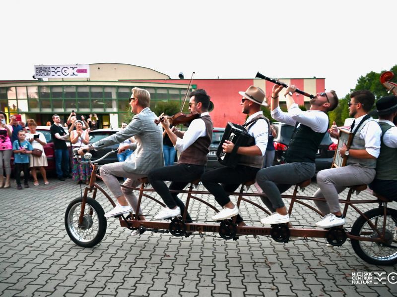 Na zdjęciu muzycy Orkiestry na Dużym Rowerze jadąc na tandemie graja na instriumentacg. Zdjęciie wykonane na parkingu MCK Tkacz