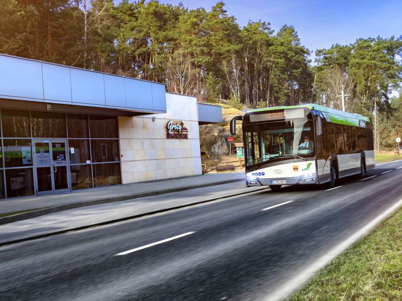 zdjęcie przedstawia autobus jadący po drodze niedleko grot nagórzyskich, widać dużo zielonych drzew budynek 