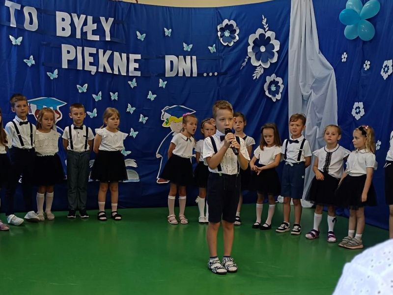 Na zdjęciu grupa przedszkolnych starszaków podczas pożegnania przedszkola. Starszaki prezentują występ artystyczny, na pierwszym planie chłopiec z mikrofonem.