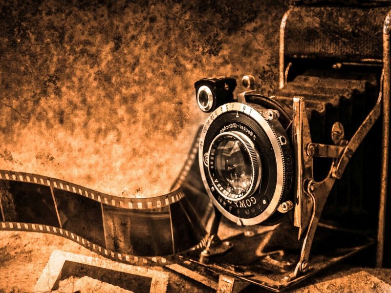 Na zdjęciu stara kamera filmowa, z której wychodzi taśma filmowa. Zdjęcie w sepii