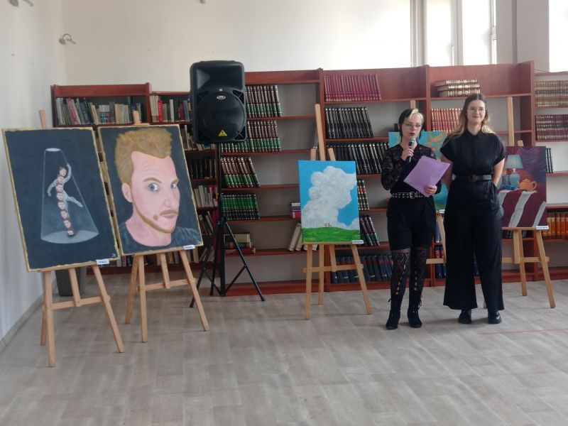 Na zdjęciu dwie kobiety, studentki Politechniki Łódzkiej wtają uczestników wernisażu prac prezentowanych w czytelni Miejskiej Biblioteki publiczbej. Wokól widac obrazy na sztalugach
