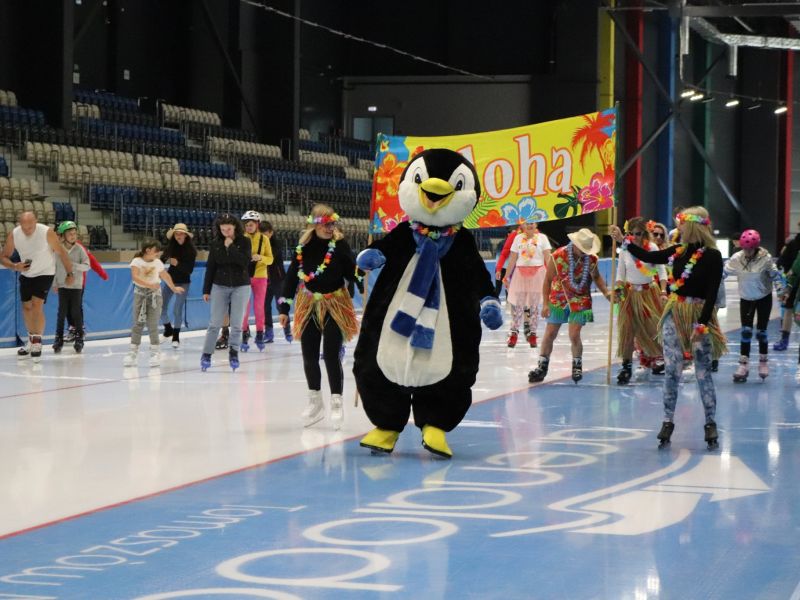 Na zdjęciu letnia ślizgawka w Areniew lodowej. Na pierwszym planie maskotka Areny - pingwin oraz dzieci w strojach tropikalnych na łyżwach