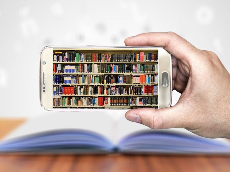 Na zdjęciu smartfon trzymany przez osobę w ręku, na ekranie smartfona widoczna biblioteka, półki z książkami