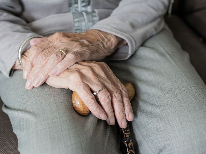 Na zdjeciu zbliżenie dłoni osoby starszej (seniora). W dłoniach senior trzyma drewnianą laskę