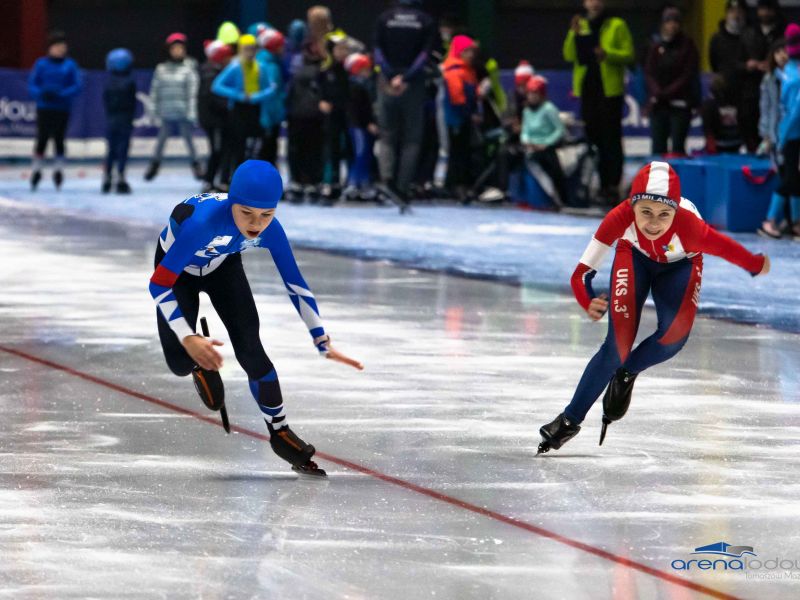 Na zdjęciu dwie łyżwiarki w kombinezonach niebieskim i czerwonym podczas wyścigu na torze lodowym. Łyżwiarki w wieku dzieci szkolnych